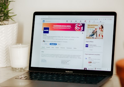 A laptop displaying a LinkedIn profile page is placed on a light-colored surface. To the left of the laptop, there is a white candle in a glass holder and a green plant in a white textured pot.