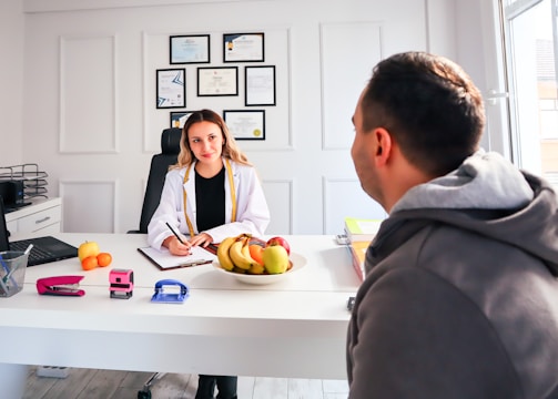 A professional consultation setting with a medical professional sitting at a desk facing a client. The room has a modern aesthetic with white walls decorated with framed certificates. The desk is organized with office supplies, a laptop, and a fruit bowl in the center.