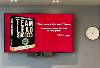 A large screen displays a book titled 'Team Lead Succeed' by Nick Fewings next to a motivational quote about teamwork attributed to the same author. The background is predominantly red. To the right of the screen, there is a white analog clock. Below the screen, a video conferencing camera is mounted.